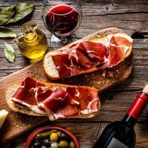 Spanish food: overhead view of two bread slices with Iberico ham also called "Bocadillo de Jamón Iberico" with red wine shot on rustic wooden table. Cheeses, olives and pickles and olive oil complete the composition. High resolution 42Mp studio digital capture taken with SONY A7rII and Zeiss Batis 40mm F2.0 CF lens